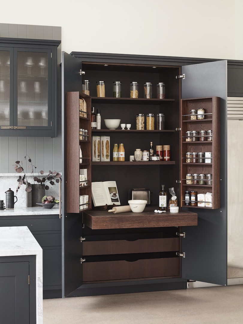 Bespoke full-height larder cupboard in charcoal grey with walnut interior shelving, door-mounted spice racks, glass storage jars, integrated cookbook stand, pull-out walnut drawers, and reeded glass wall cabinets visible beyond