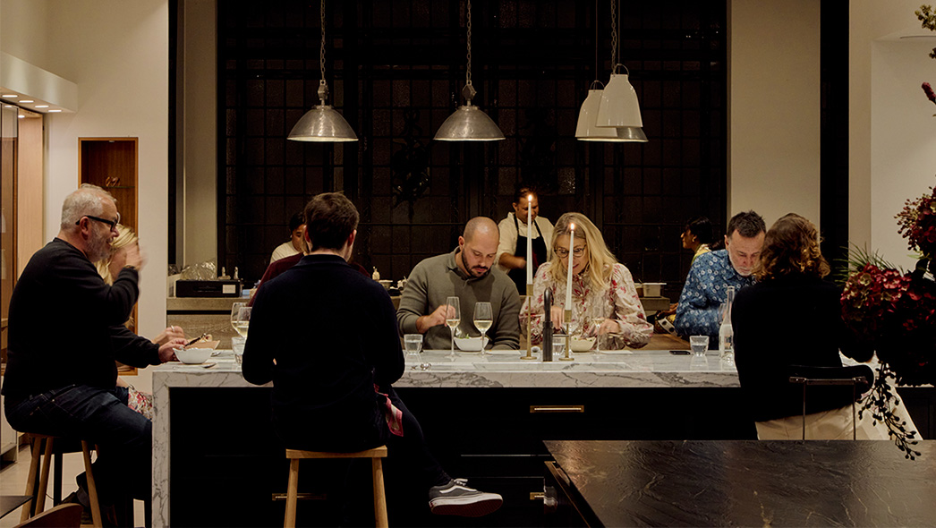 Evening dining event at Roundhouse showroom with guests seated around white marble island, industrial metal pendant lights, floor-to-ceiling black-framed glazing, and warm candlelit atmosphere