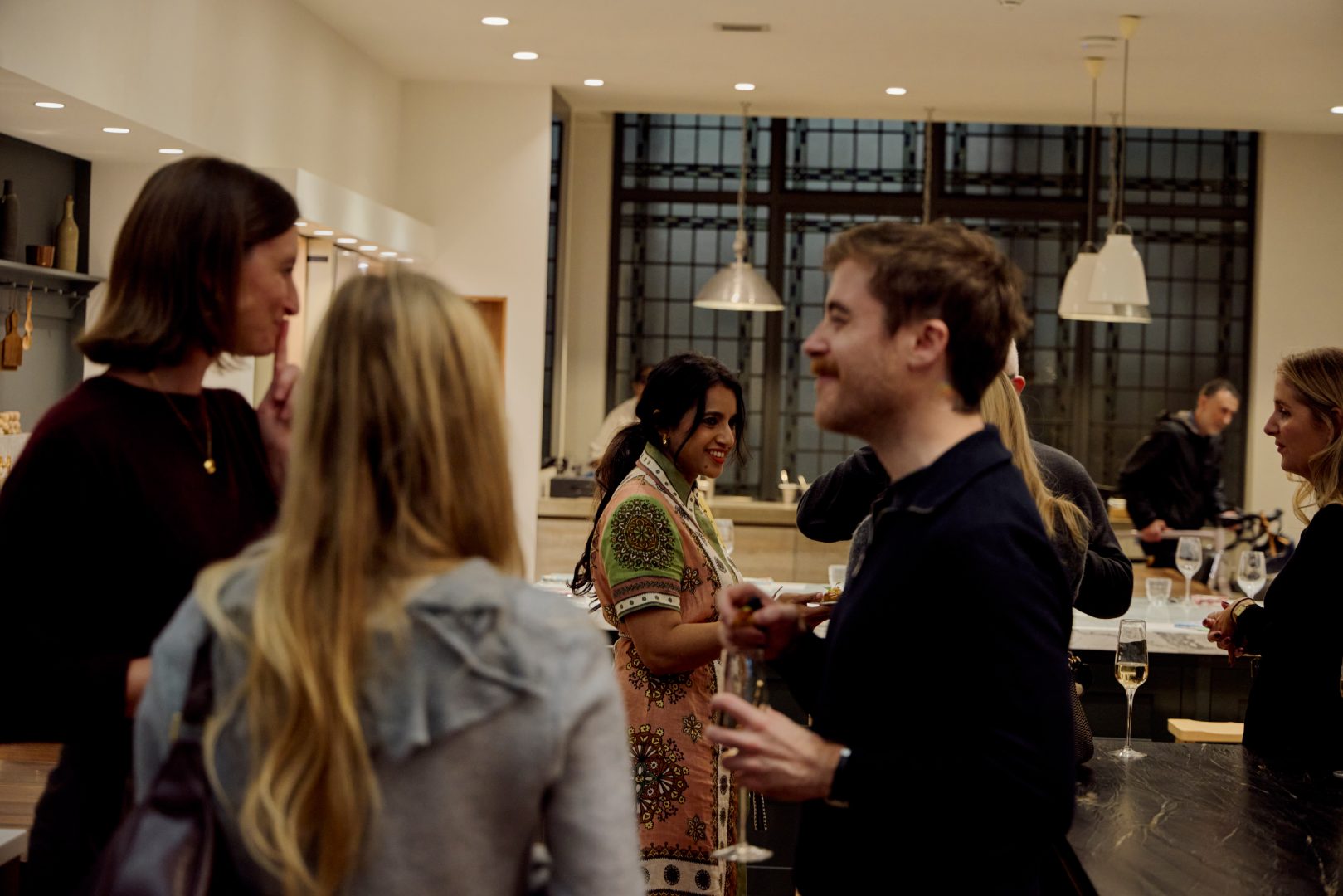 Networking event at Roundhouse showroom with guests mingling around kitchen island, black-framed Crittall-style glazing, white pendant lights, and illuminated display cabinetry in background