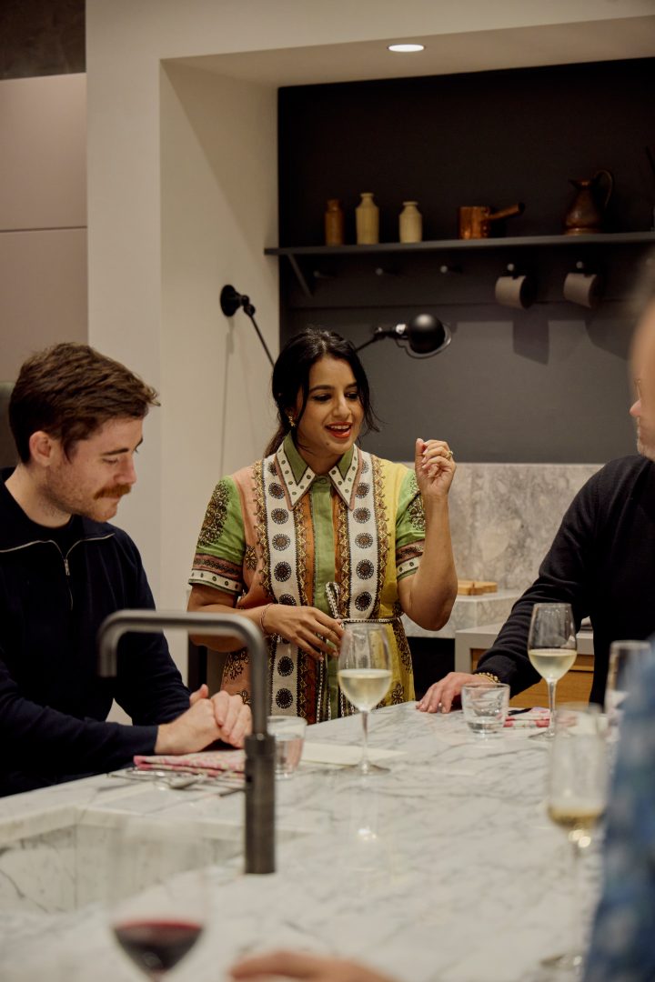 Cooking workshop at Roundhouse showroom with chef demonstrating culinary techniques at white marble island, guests seated with wine glasses, dark feature wall with floating shelf displaying ceramics, and matt black tap visible