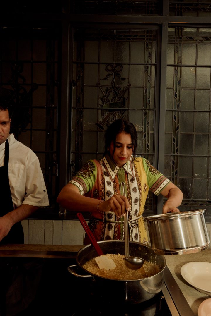 Cooking demonstration at Roundhouse showroom with chef preparing dish in large pan, industrial steel-framed glazing backdrop, stainless steel cookware, and intimate event atmosphere