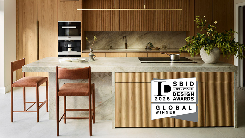 Award-winning Roundhouse bespoke kitchen featuring vertical oak veneer cabinetry, pale marble island worktop with fluted wood panelling, and bronze tap with integrated hob – SBID International Design Awards 2025 Global Winner