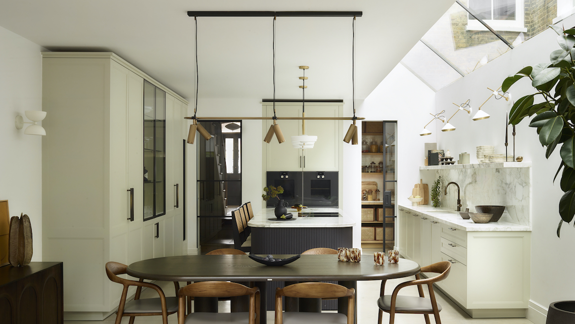Contemporary kitchen extension with vaulted ceiling and skylight, pale sage shaker cabinetry, black-framed Crittall-style glazed partitions, white marble splashback, dark charcoal island with fluted base, round walnut dining table with tan leather chairs, brass cone pendant lights, and architectural rooflight flooding space with natural light
