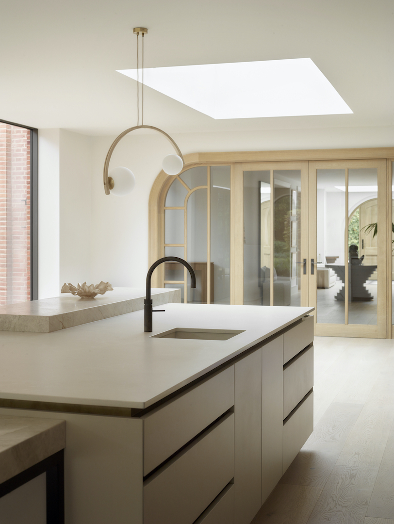 Sculptural minimalist kitchen with pale stone-effect island, matt black swan-neck tap, natural oak arched glazed doors, statement brass ring pendant light, rectangular ceiling void skylight, and white oak flooring