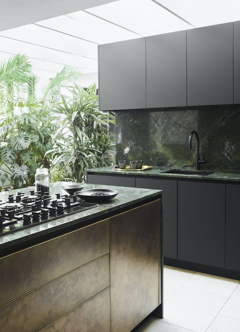 Dramatic dark grey kitchen with deep green marble worktops and splashback, antiqued brass island with brass rail details, matt black tap, flush-mounted black hob, tropical plants in conservatory beyond, and white tile flooring