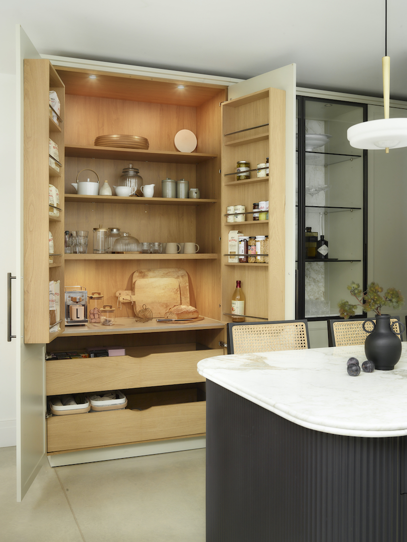 Oak larder cupboard with integrated lighting revealing organised storage shelves, pull-out worktop and deep drawers beside fluted kitchen island with marble top
