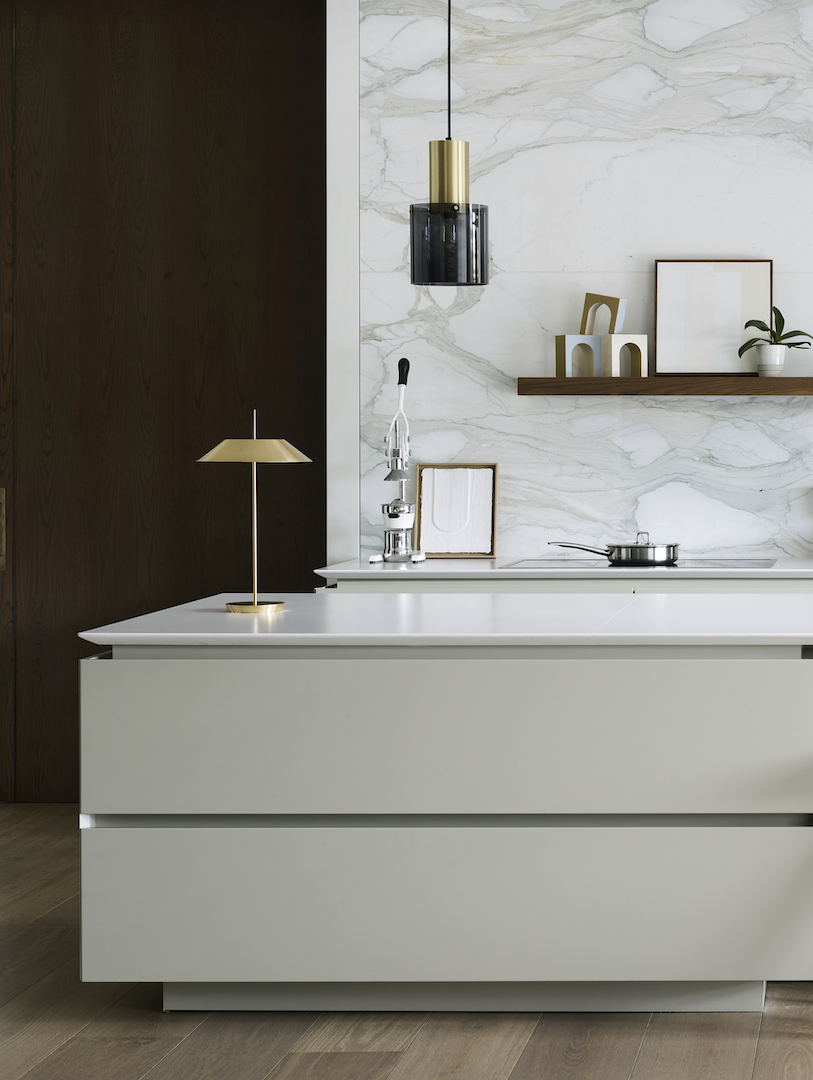Sophisticated minimalist kitchen with white handleless island, Calacatta marble splashback wall, walnut floating shelf, brass table lamp and pendant lights, dark oak timber wall cladding, and curated sculptural objects