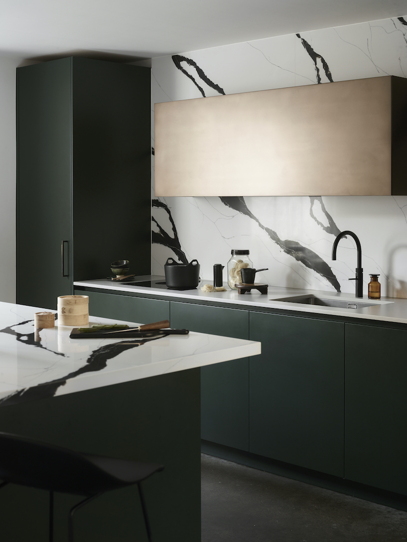 Sophisticated forest green handleless kitchen with dramatic bookmatched white marble worktops and splashback featuring bold black veining, integrated pale timber feature panel, matt black swan-neck tap, and moody atmospheric lighting