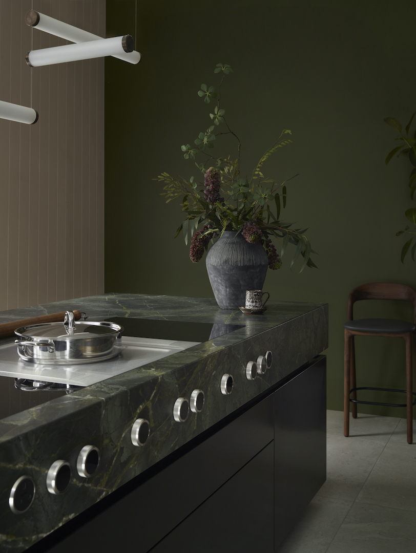 Dark green marble worktop with circular brass tap details and chrome hob controls against sage green painted walls in luxury bespoke kitchen