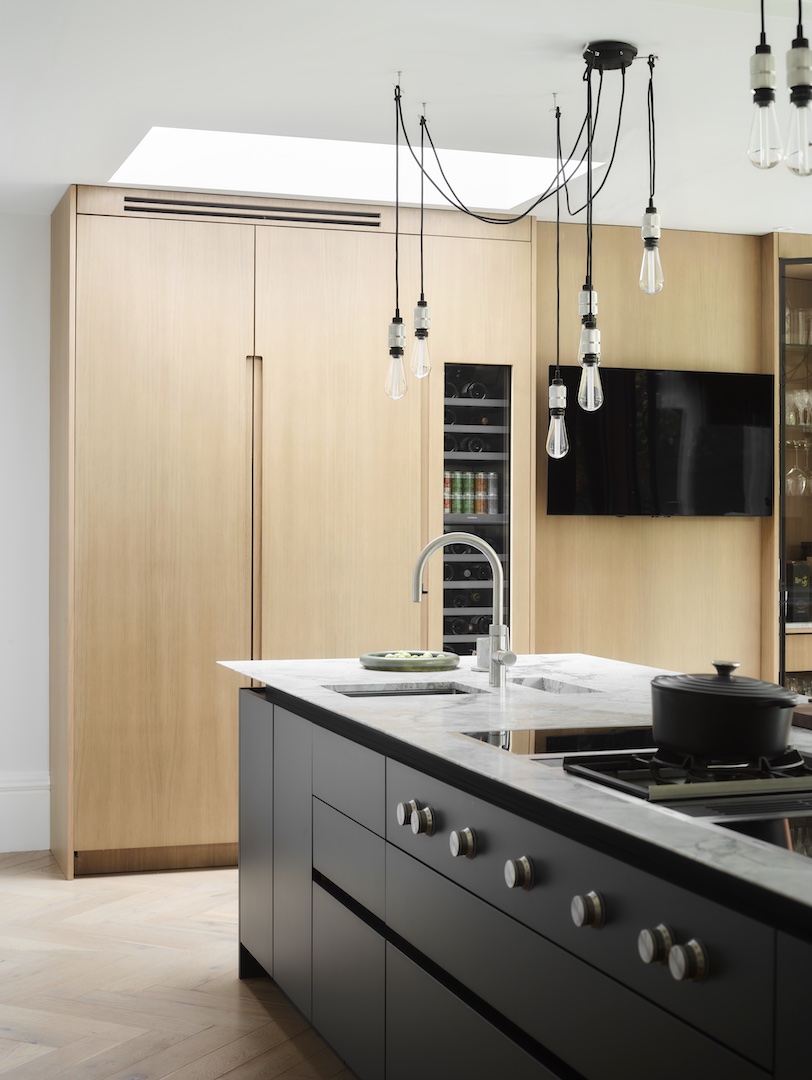 Contemporary kitchen with natural oak wall cabinetry, charcoal grey island with white marble worktop, integrated wine cooler and fridge columns, industrial Edison bulb spider chandelier, brushed steel tap, and herringbone oak flooring