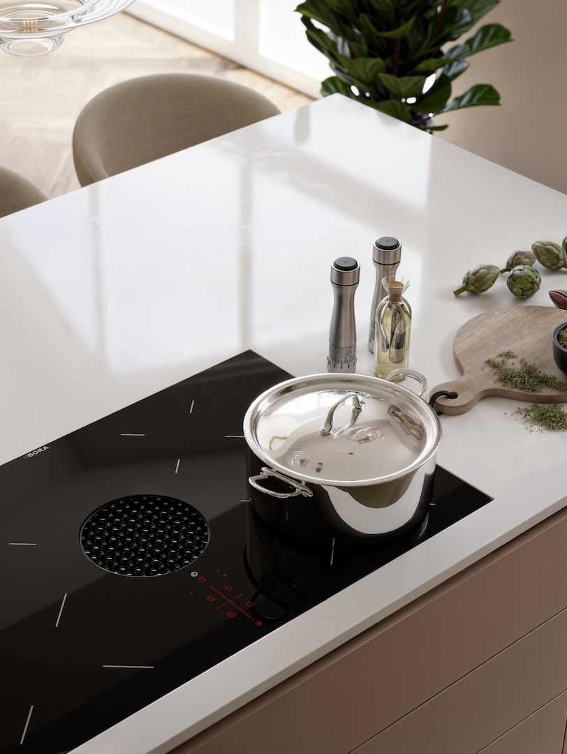 Minimalist kitchen detail featuring flush-mounted black induction hob with integrated extraction zone, seamless white composite worktop, stainless steel cookware, oak cabinetry, and curated cooking accessories including olive oil and herbs