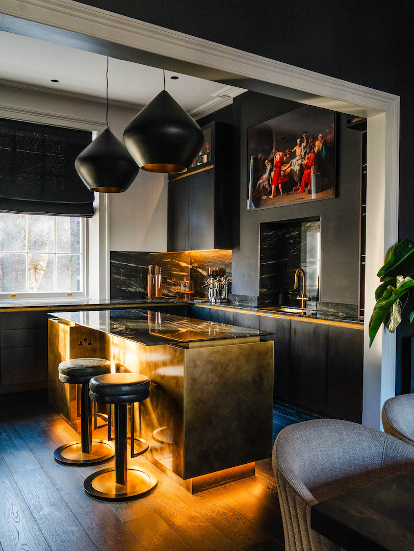 Dramatic dark kitchen with charcoal cabinetry, statement brass-clad island with black marble worktop, gold-veined black marble splashback with LED lighting, brass tap, black dome pendant lights, black leather bar stools, period sash window, classical artwork, and dark oak flooring
