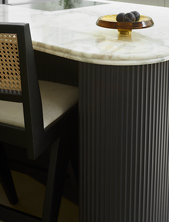 Bespoke kitchen island with fluted black cabinetry and white marble worktop, cane-back dining chair, brass cake stand with fresh fruit