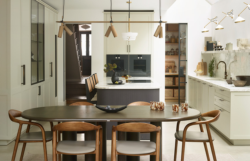 Bespoke luxury kitchen with sage green shaker cabinetry, fluted charcoal island, marble worktops, brass pendant lighting, and integrated dining area with curved walnut chairs