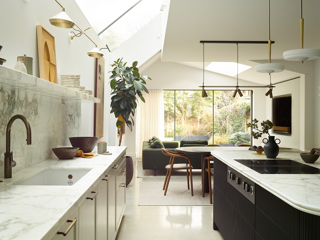 Open-plan bespoke kitchen with dramatic rooflight, marble splashback and worktops, fluted island with integrated hob, bronze tap, and garden views through floor-to-ceiling glazing