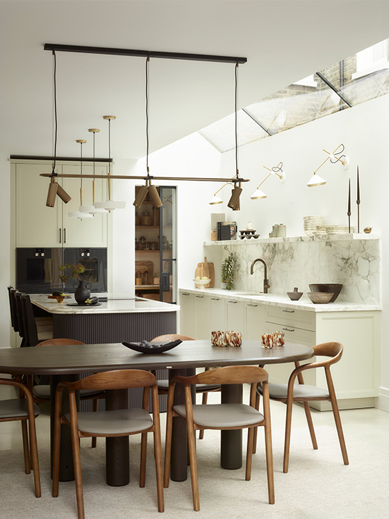 Contemporary kitchen-dining space featuring sage cabinetry, charcoal fluted island, brass architectural pendants, marble worktop with bronze tap, and oval dining table with walnut chairs