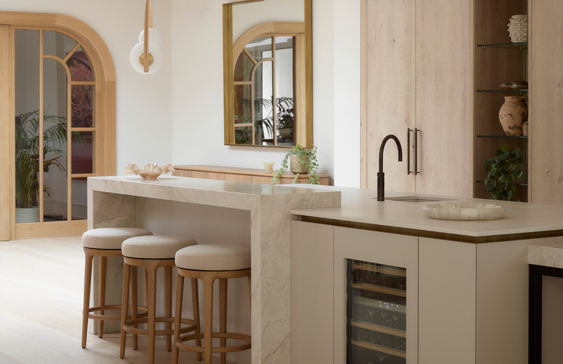 Mediterranean-inspired kitchen with limewash-textured pale cabinetry, arched brass-framed mirror openings, oak bar stools with cream linen seats, integrated wine cooler, and travertine-effect composite worktop