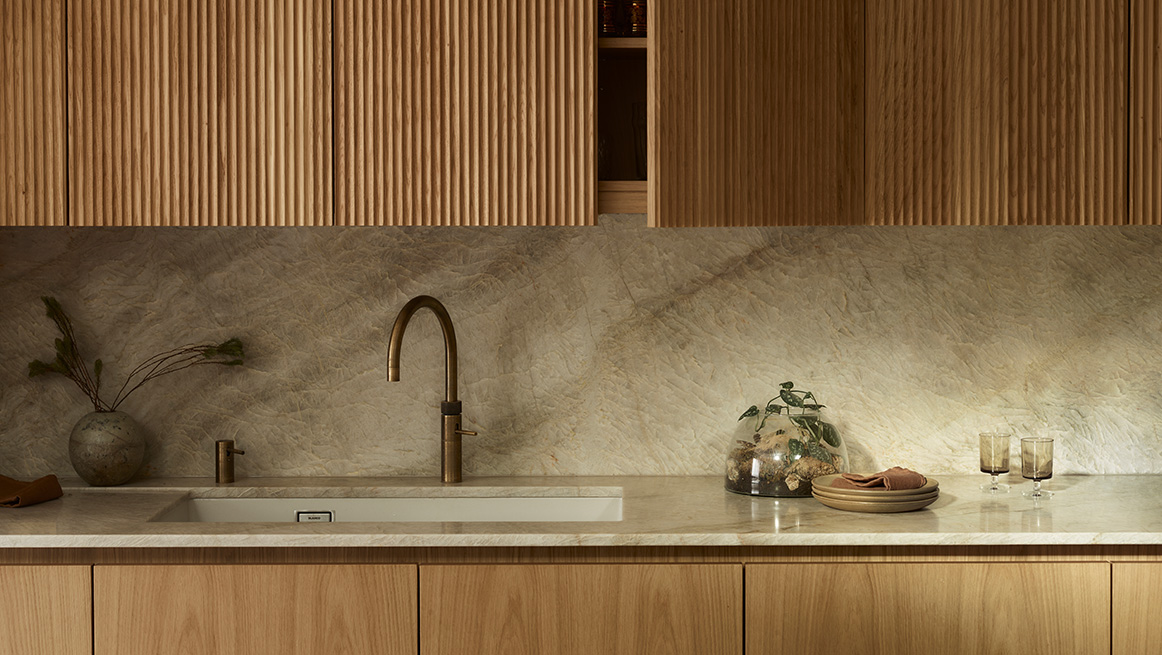 Natural oak handleless kitchen with vertical fluted timber wall panels, limestone splashback, and brushed brass tap in contemporary British interior