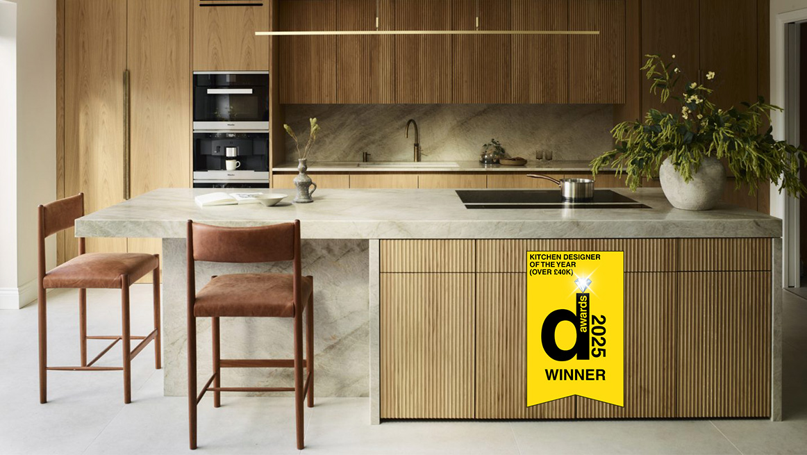 Bespoke luxury kitchen with vertical oak fluted island cabinetry, pale green marble worktop, and integrated sink, featuring Design Awards 2025 Kitchen Designer of the Year winner badge, mid-century leather bar stools, and linear pendant lighting
