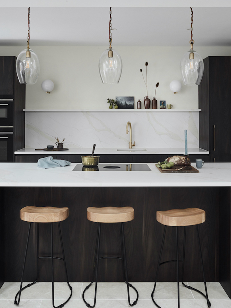 Sophisticated dark stained oak kitchen with handleless cabinetry, white marble island worktop and splashback, brass tap and handles, clear glass pendant lights with brass fittings, natural oak bar stools with black metal legs, and white floating shelf