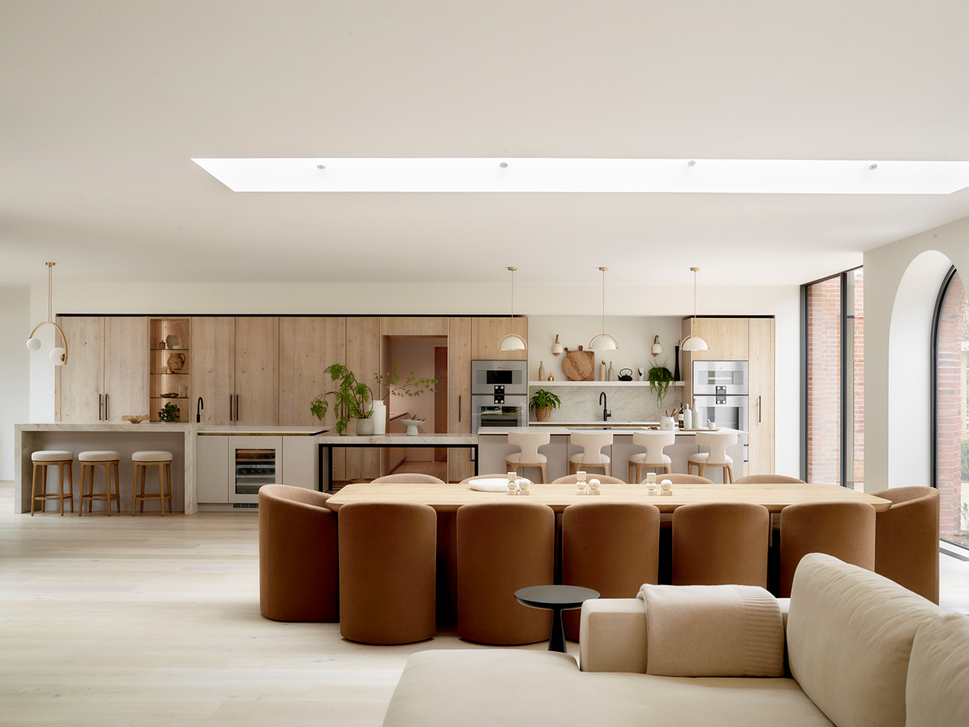 Expansive open-plan kitchen and dining space with natural oak wall-to-wall cabinetry, white painted islands, oval dining table with camel velvet chairs, white upholstered bar stools, brass pendant lights, integrated wine cooler, and floor-to-ceiling glazing
