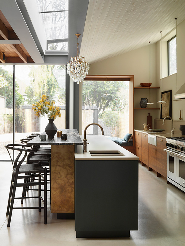 Dramatic kitchen extension with linear rooflight, vaulted tongue-and-groove ceiling, natural oak shaker cabinetry, dark marble waterfall island with brass tap, black wishbone dining chairs, crystal chandelier, walnut floating shelves, and floor-to-ceiling garden views