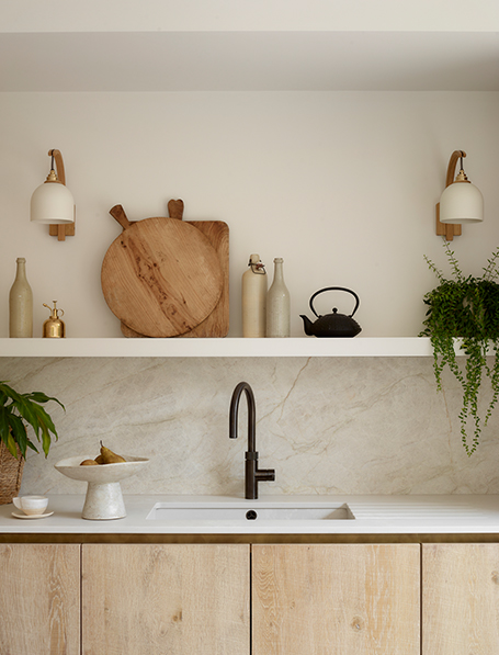 Minimalist kitchen detail with pale oak base cabinets, white marble worktop and splashback, white floating shelves displaying wooden serving boards and ceramics, matt black tap, and brass wall sconces