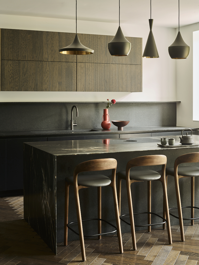Modern kitchen island with textured dark oak cladding, honed stone worktop, Tom Dixon Beat pendants and fluted oak wall cabinetry with minimalist grey splashback