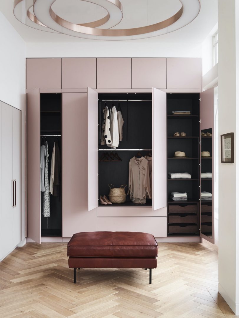 Bespoke blush pink fitted wardrobe system with open doors revealing organised interior storage, black shelving, hanging rails, and herringbone oak flooring with tan leather ottoman