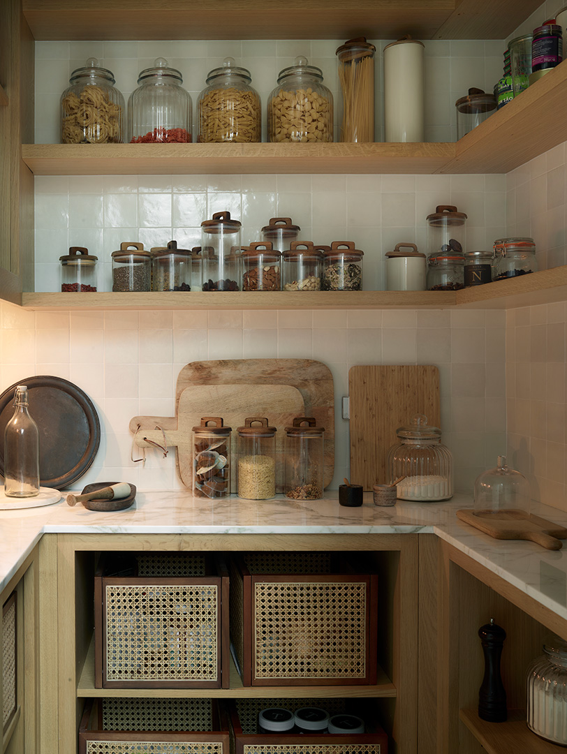Bespoke pale oak open shelving with white metro tiles, displaying glass storage jars with wooden lids and chopping boards, integrated beneath-shelf lighting, sage green cabinetry with cane webbing panel detail below marble worktop