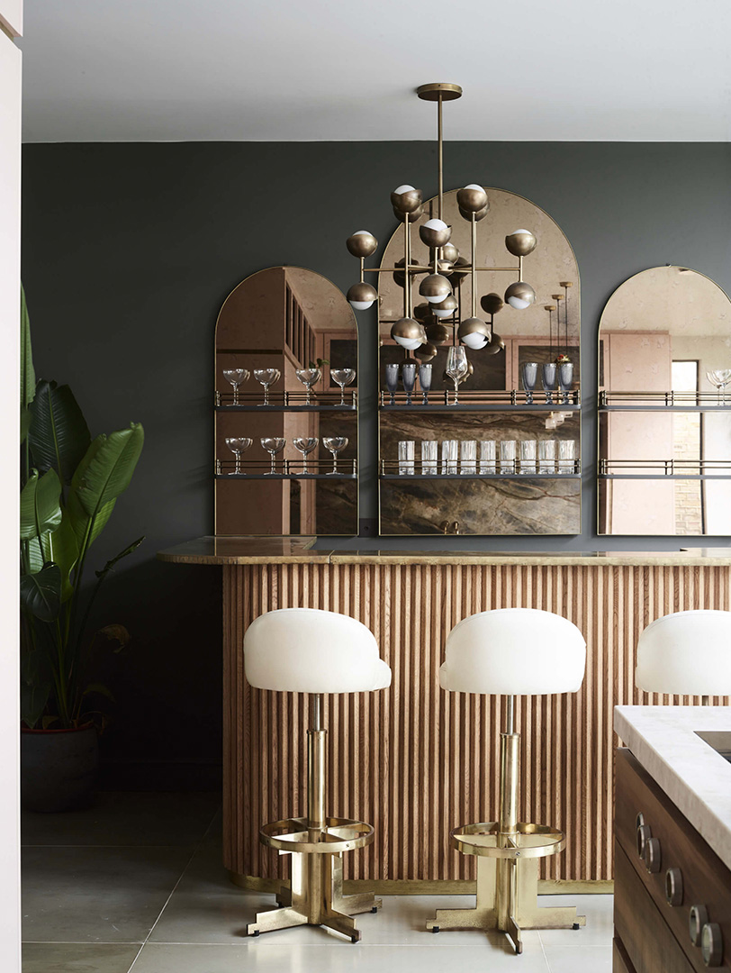 Luxury Roundhouse kitchen bar with vertical fluted oak cabinetry, bronze-framed arched mirror cabinets displaying glassware, antique brass Sputnik chandelier, and bespoke brass bar stools with cream bouclé upholstery against charcoal grey walls
