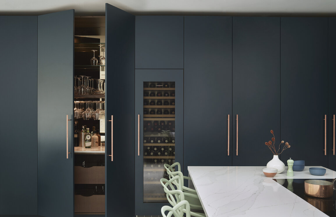 Bespoke deep teal handleless kitchen with integrated glassware storage featuring copper bar handles, built-in wine cooler, and white marble island with sage green dining chairs