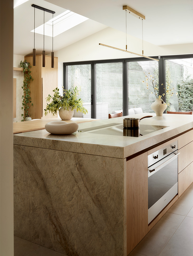 Contemporary pale oak kitchen island with book-matched marble waterfall edge, vaulted ceiling with skylight, bronze pendant lighting, black aluminium crittal-style doors overlooking landscaped courtyard, ceramic display pieces