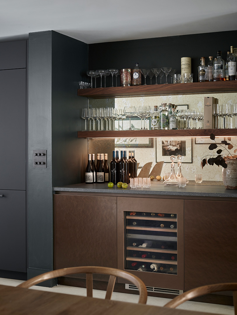 Bespoke dark grey kitchen with walnut open shelving bar area, integrated wine cooler, under-shelf LED lighting, glassware display, marble splashback with bronze tap, entertaining zone design