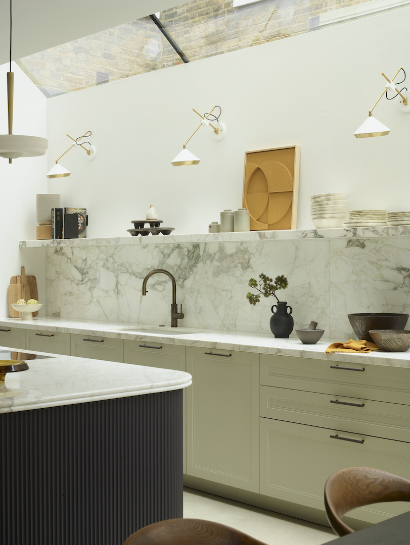 Sage green handleless kitchen cabinetry with full-height Carrara marble splashback, brass adjustable wall lights, and curved fluted island detail beneath exposed brick skylight