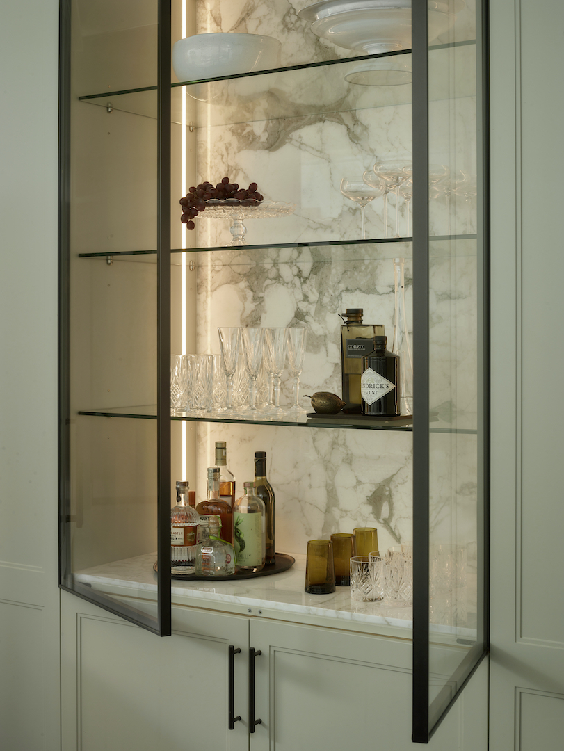 Bespoke illuminated drinks cabinet with bronze-framed glass doors, marble back panel, and floating glass shelves displaying crystal glassware and spirits