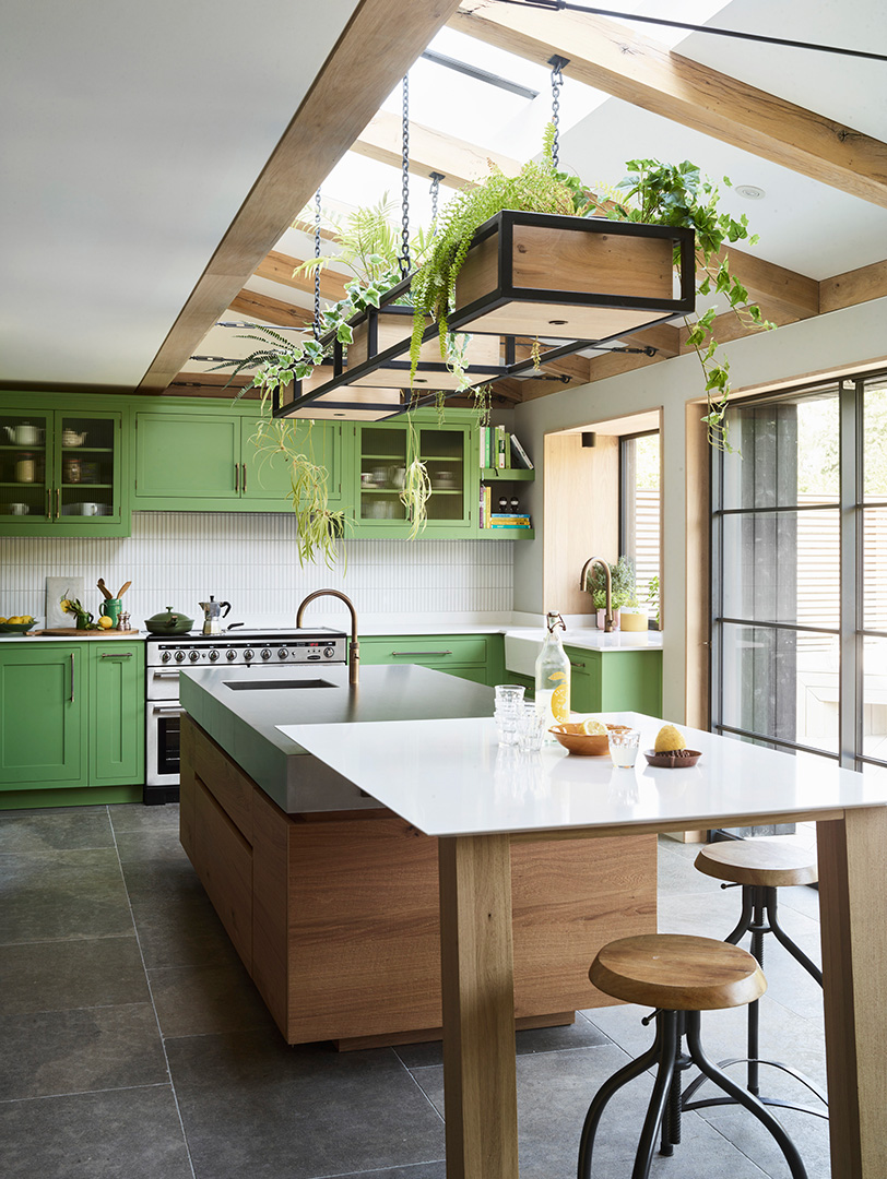 Sage green Shaker kitchen with exposed oak beams, suspended plant shelving, two-tone oak island with integrated dining table, and slate flooring in vaulted extension