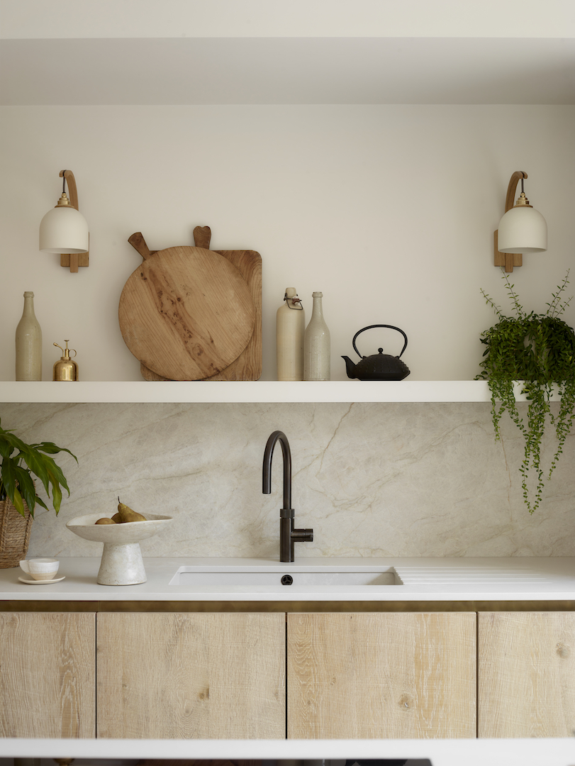 Natural oak handleless cabinetry with white marble splashback, bronze tap, floating shelf with artisan ceramics, and oak and brass wall sconces