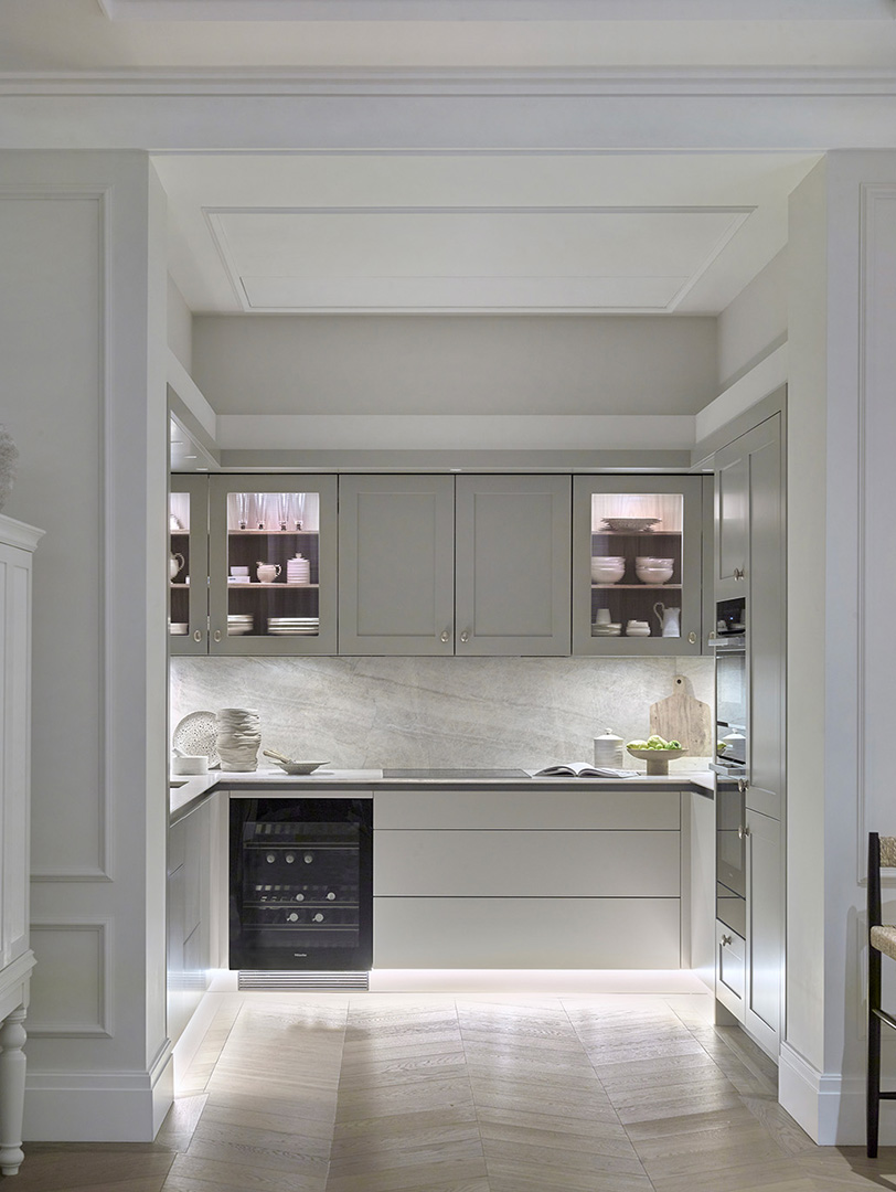 Luxury bespoke butler's pantry with pale grey Shaker cabinetry, illuminated glass-fronted display cupboards, white marble splashback, integrated wine cooler, and architectural coffered ceiling detail