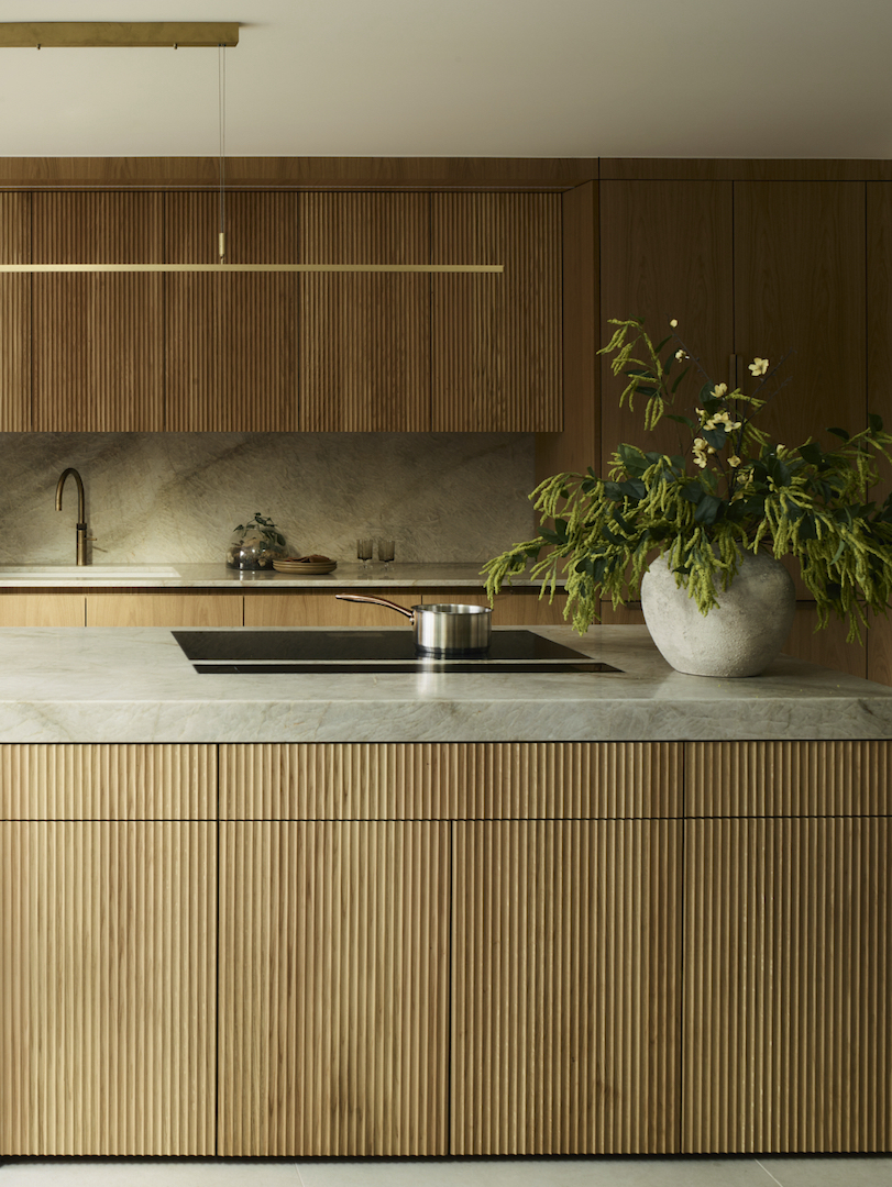Contemporary fluted oak kitchen island with grey marble worktop, flush-mounted induction hob, brass tap, and textured stone splashback in earth-toned luxury kitchen