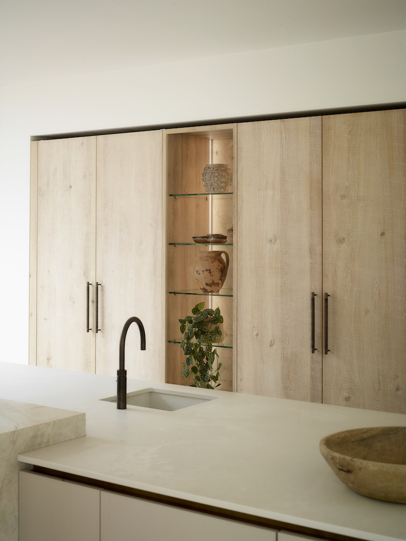Minimalist bespoke kitchen with light oak wall units, integrated glass shelving displaying ceramics, cream stone worktop, and matte black tap with undermounted sink