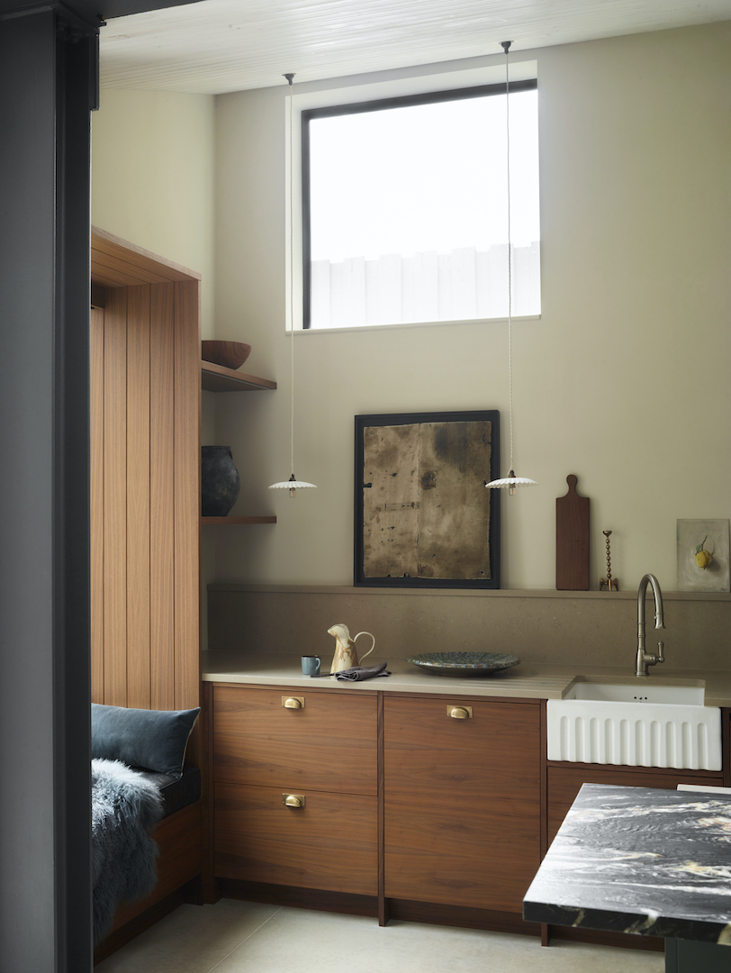 Mid-century inspired walnut kitchen featuring brass cup handles, fluted Belfast sink, open timber shelving with artisan ceramics, and statement pendant lighting beneath skylight