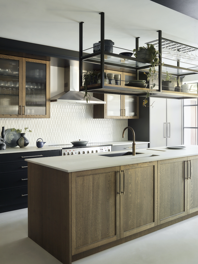 Architectural kitchen with suspended black metal shelving system, smoked oak island, fluted glass cabinet doors, white geometric splashback tiles, and professional range cooker