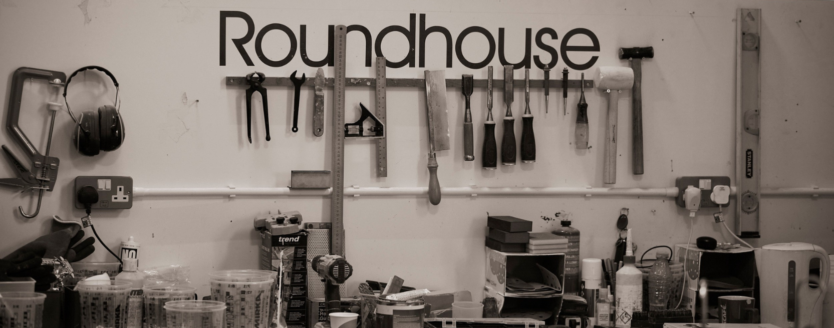 A British workshop with tools hanging on a wall, labeled "Roundhouse," above a workbench cluttered with various items.