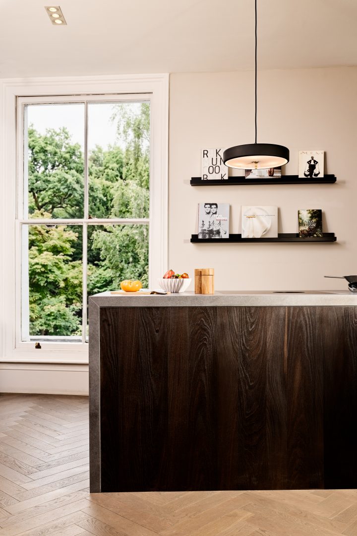 Contemporary kitchen display featuring a dark timber island with a stone worktop, integrated invisible induction technology, open shelving and large sash windows overlooking greenery at the Roundhouse + Day True Wimbledon showroom.