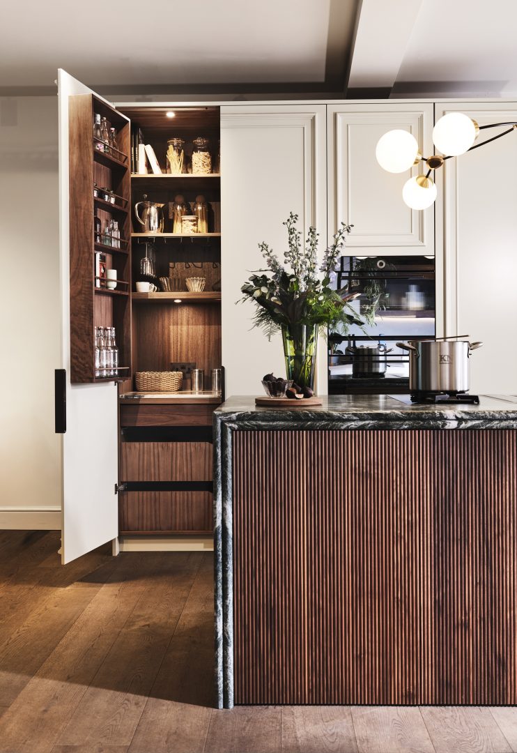 Luxury kitchen display with a fluted walnut island wrapped in dramatic double bullnose stone worktop, illuminated walnut pantry storage, shaker-style cabinetry and bespoke joinery at the Roundhouse + Day True Wimbledon showroom.