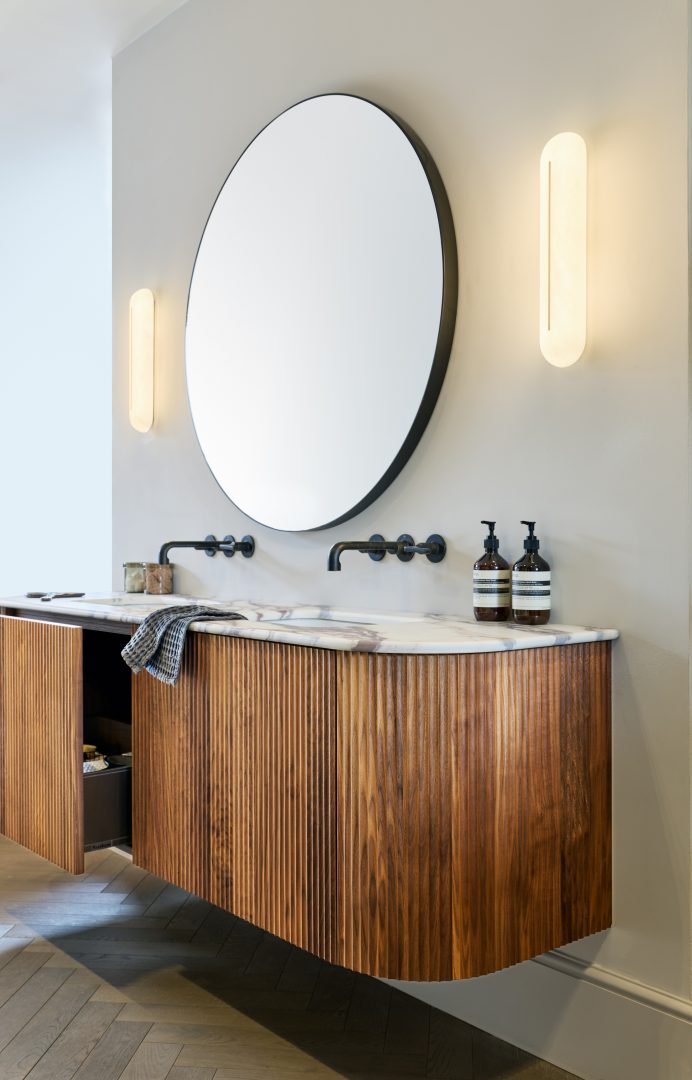 Floating fluted walnut bespoke bathroom vanity with a Calacatta marble worktop, integrated basin, black wall-mounted taps, circular mirror and contemporary wall lights at the Roundhouse + Day True Wimbledon showroom.