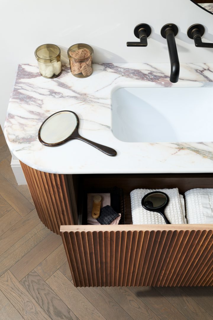 Detail of a Calacatta marble bathroom worktop with integrated basin, black wall-mounted tapware and bespoke fluted walnut cabinetry at the Roundhouse + Day True Wimbledon showroom.