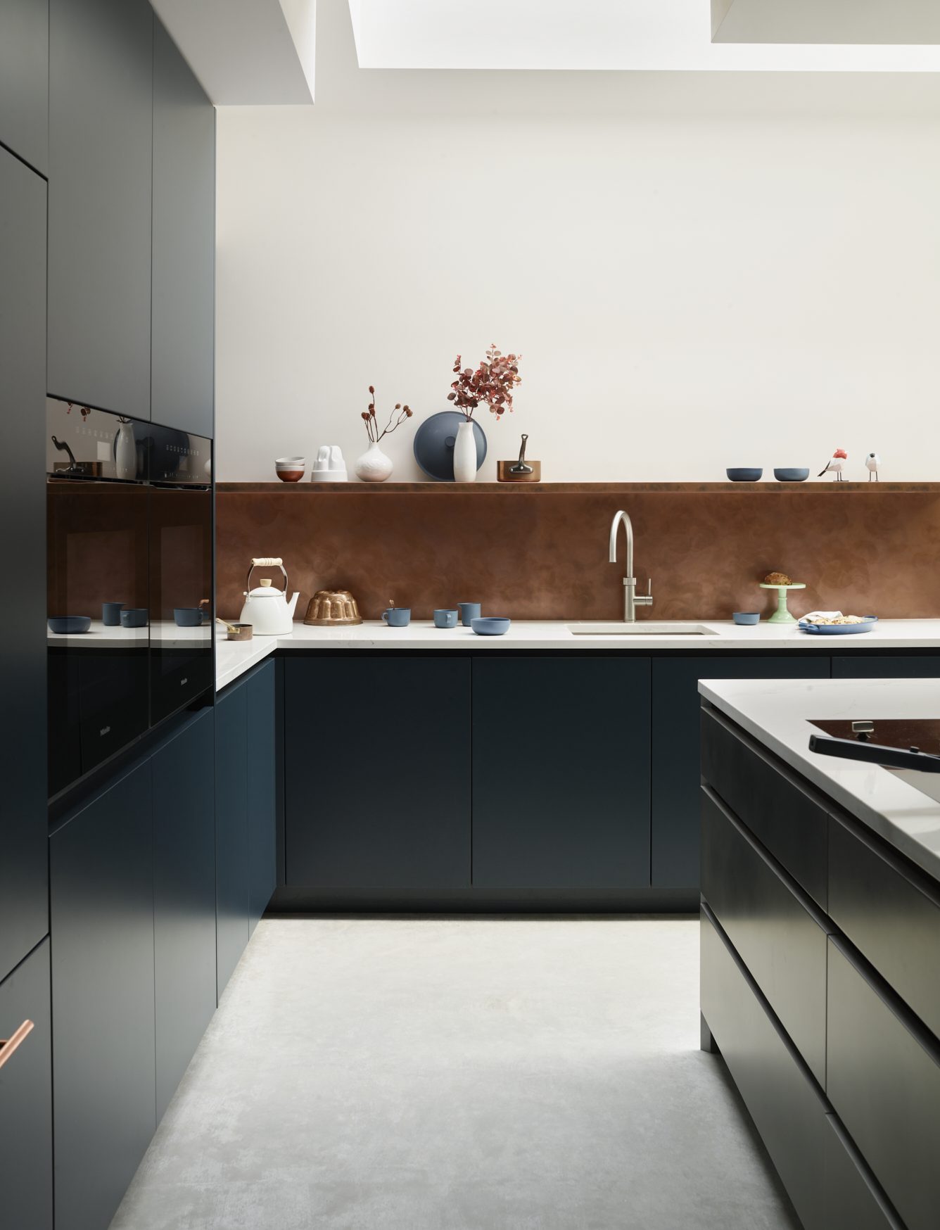Contemporary bespoke kitchen featuring dark blue handleless cabinetry, white worktops and a warm copper-toned metallic splashback. Natural light pours through a large rooflight, highlighting minimalist styling, integrated appliances and clean architectural lines.