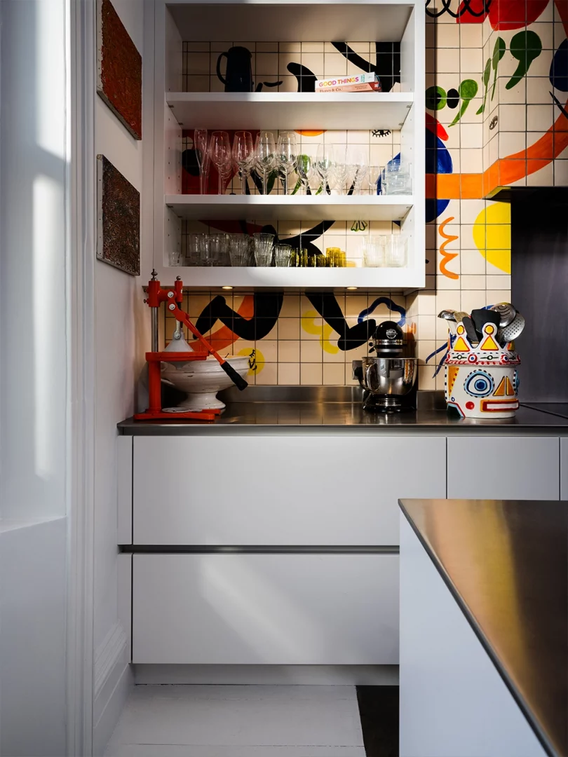 Open shelving and white bespoke cabinetry in Yotam Ottolenghi's Roundhouse kitchen, set against a hand-painted tiled splashback with colourful abstract artwork and stainless steel worktops.